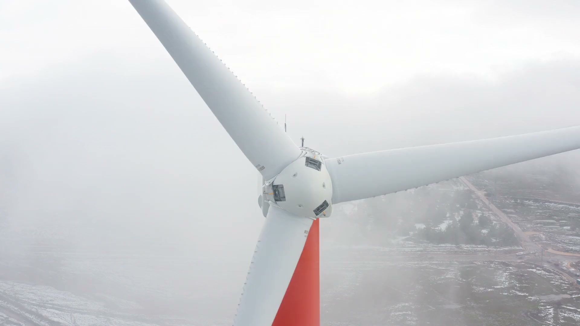 The hub and blades of a wind turbine in fog, seen from the air, its red tower dropping away below