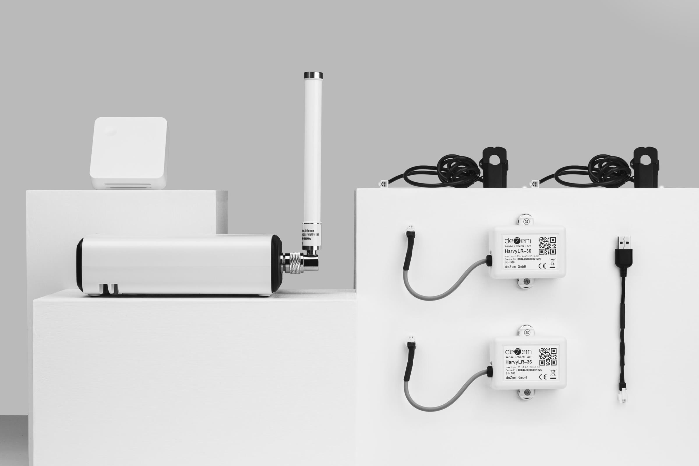 DEZEM's LoRaWAN starter kit on white plinths: a gateway with its antenna, a room sensor and two wireless meters with their current clamps