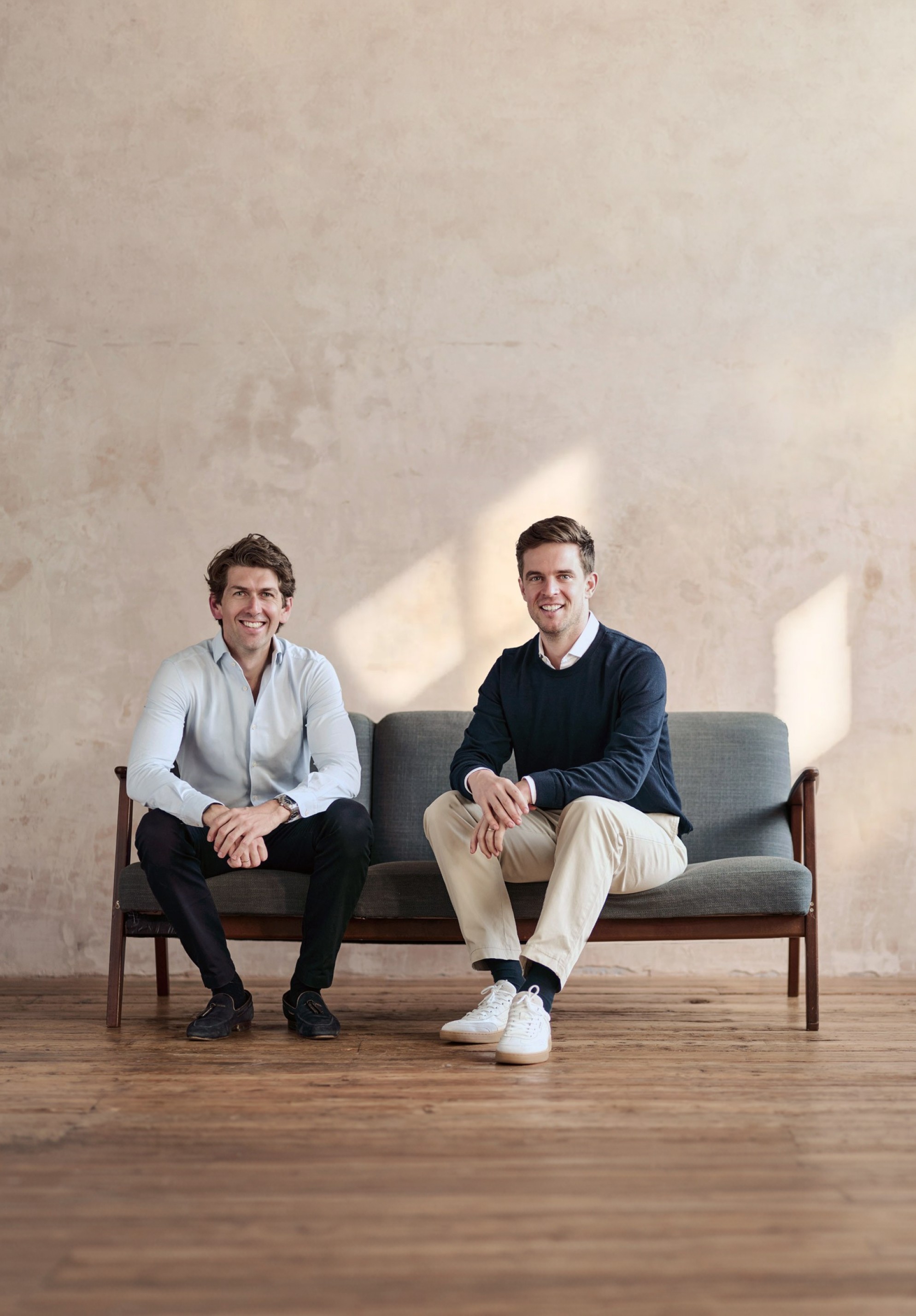 The two founders sitting on a sofa in front of a plastered wall, laughing