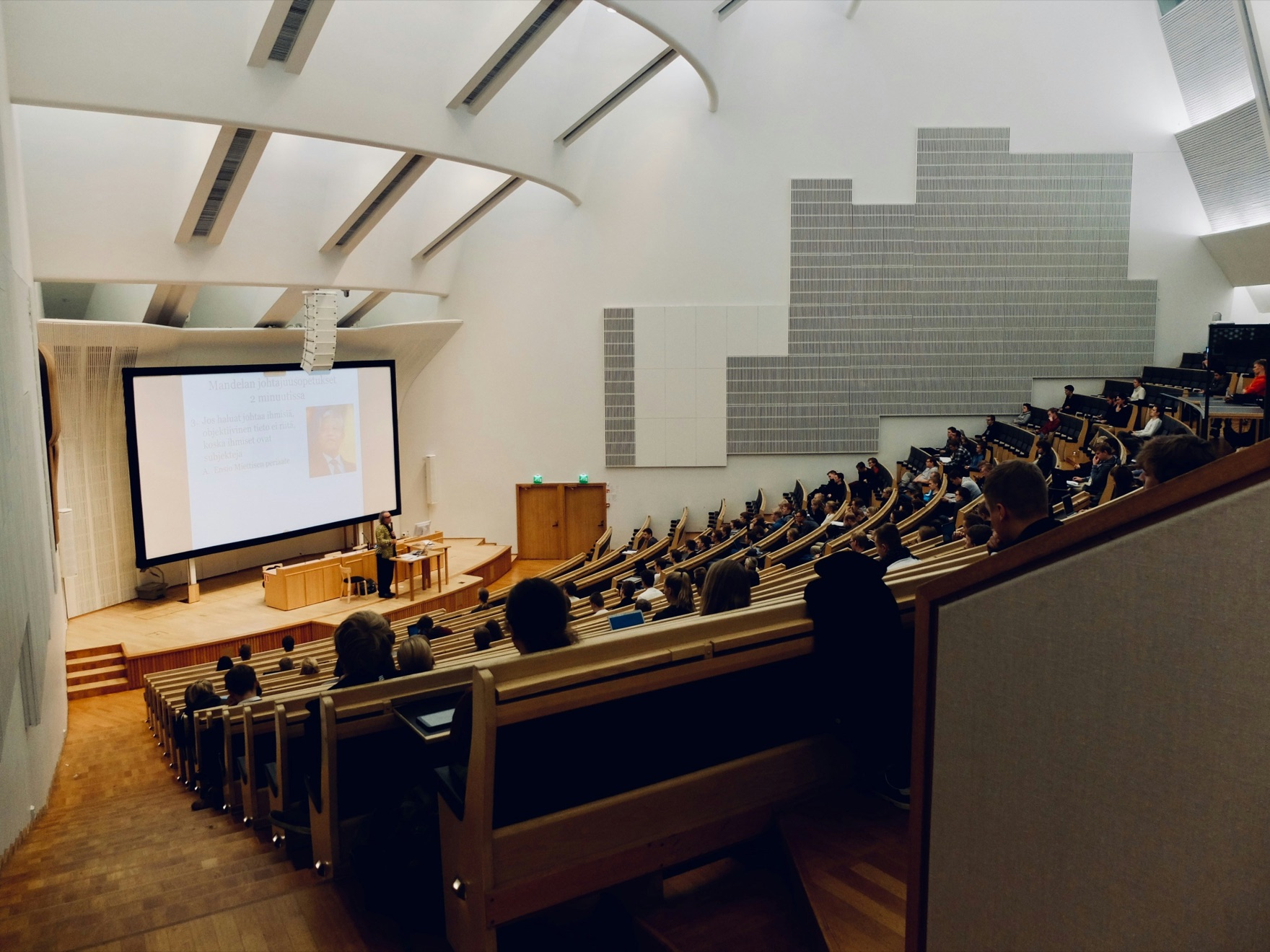 A lecture hall seen from the back rows, the audience facing a lecturer beside a large projection screen